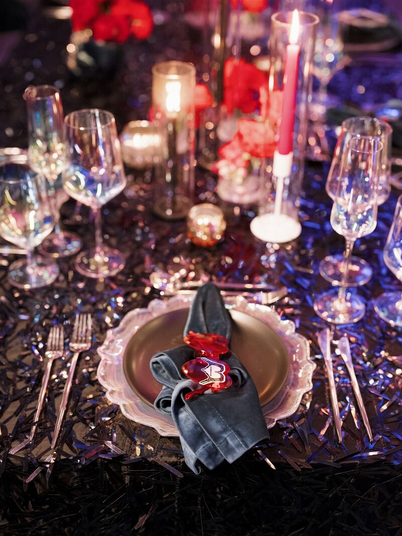 aerial view of a modern gothic tablescape on a black sequin tablecloth with iridescent glassware, silver flatware, iridescent charger plate, black dinner plate with a teal velvet napkin on top with heart shaped hot pink transparent glasses and skull heart sticker placecard