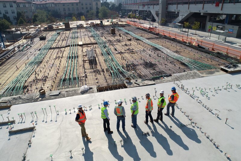 Group ofEngineer workers and supervisors wearing safety vests and hard hats stand on a concrete slab, overlooking a large construction site with rebar and green pipes laid out for the foundation.