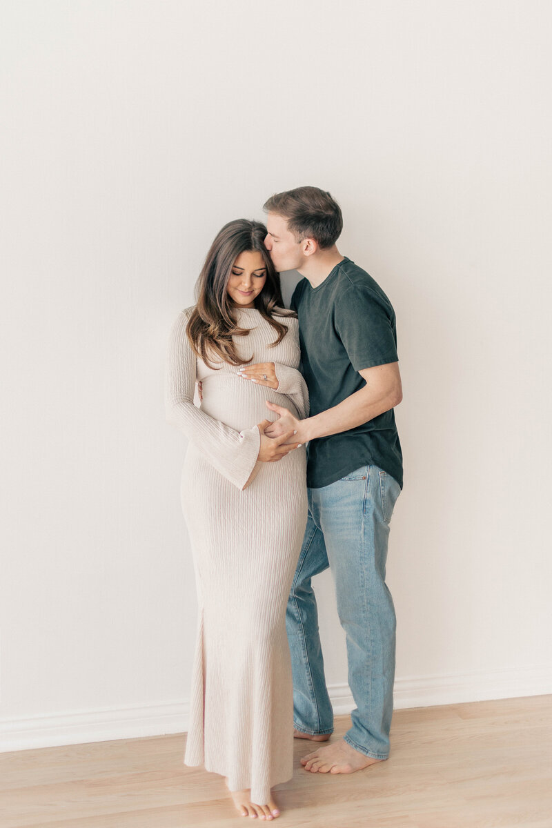 Pregnant woman in a cream dress holding her belly while her partner embraces her from behind, both barefoot against a clean studio backdrop in Kansas City.