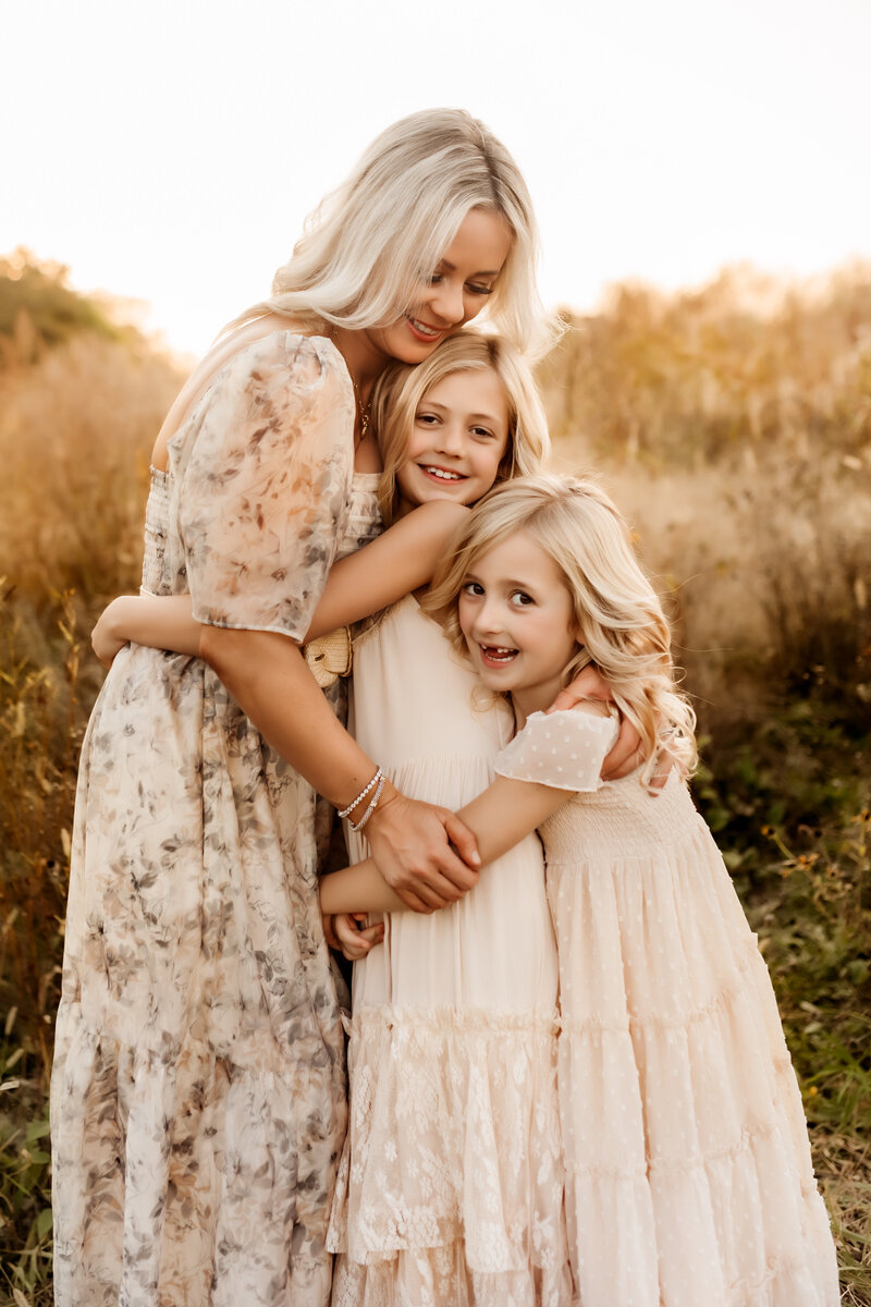 Mom smiles down at her two girls as they hug each other in beautiful dresses in Family Photoshoot in  Kansas City.