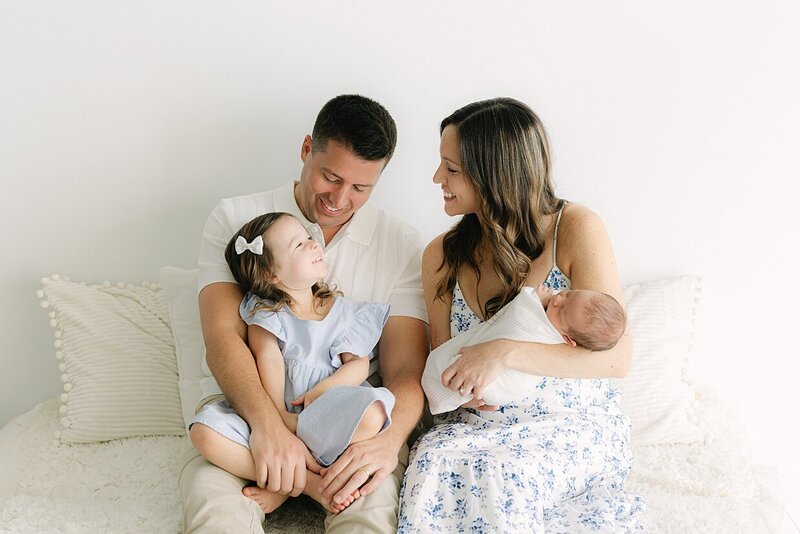 Toddler girl in blue dress with white bow sits on her dad's lap smiling at her mother in a blue floral dress holding her newborn brother during their Indianapolis studio newborn photography session