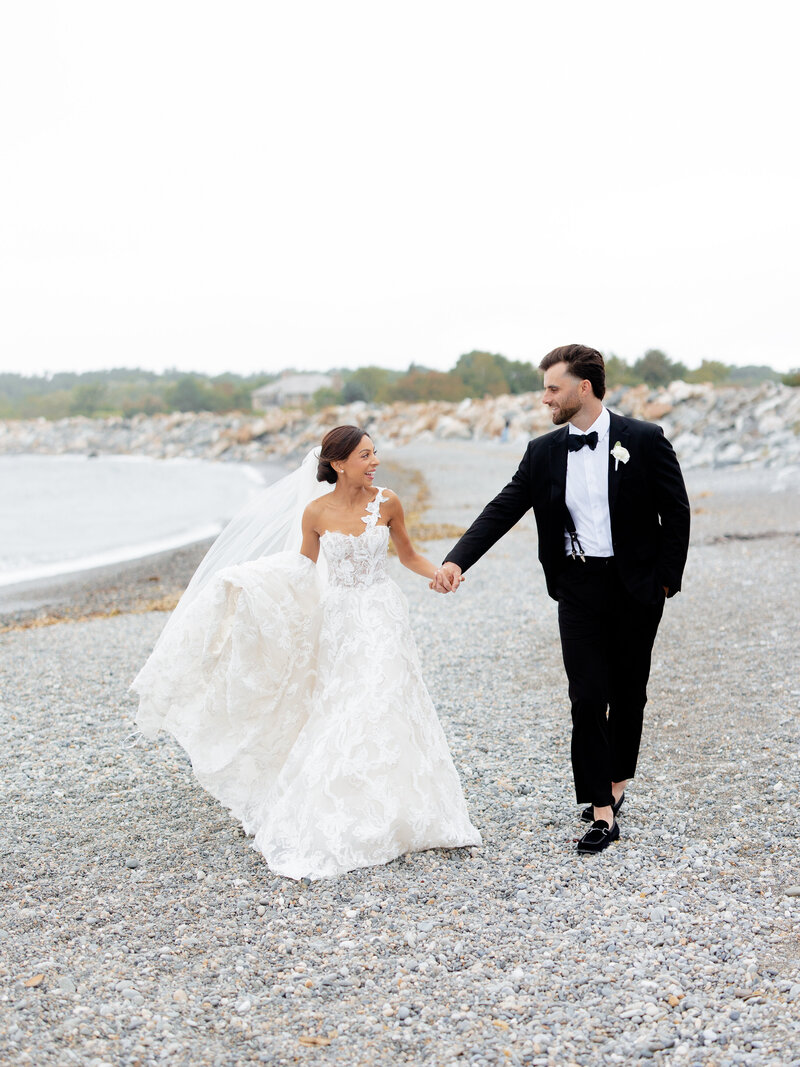 Bride and groom walk hand in hand on a rocky beach.