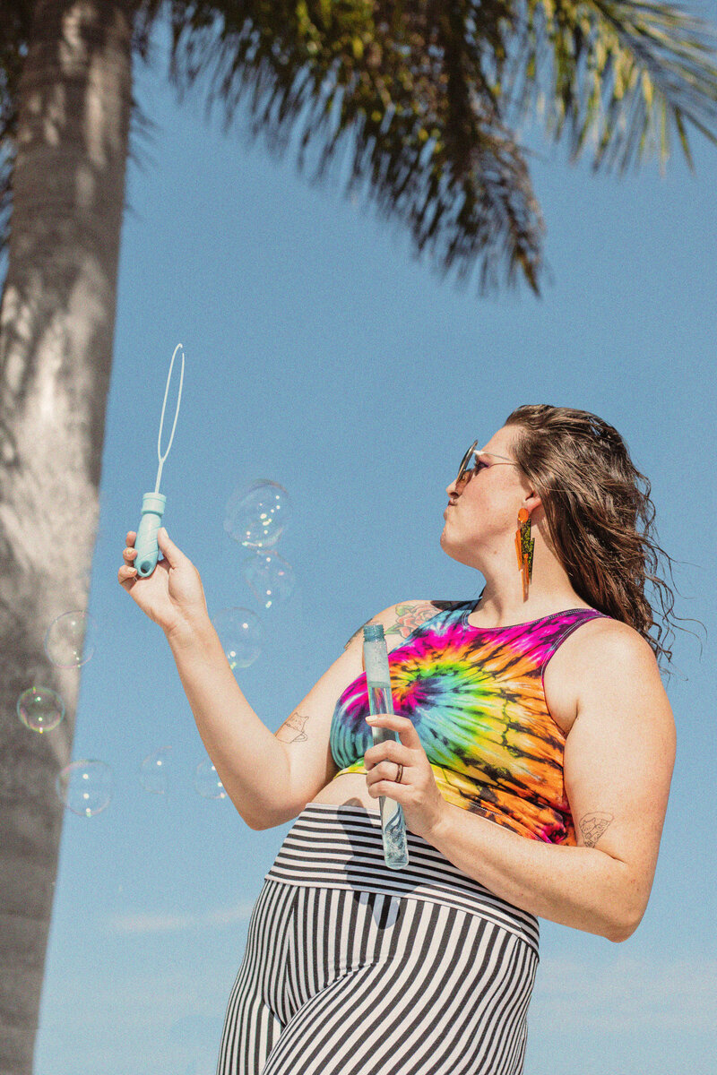 Model wearing lifestyle clothing poses in front of palm tree while blowing bubbles