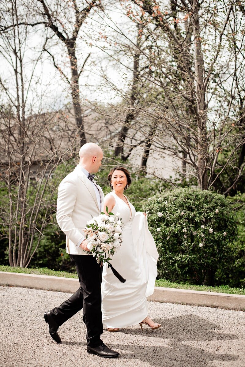 The bride and groom go for a walk at the Noah Liff Opera Center. The groom is wearing black pants and a white tuxedo jacket. He is carrying the bride's large white bouquet with long black ribbons as he looks over at her. The bride is wearing a fitted sleeveless sheath dress  with a very long train and a plunging neckline. The bride holds her train as she walks.