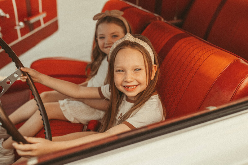 Two young girls smiling in a vintage car with red interior.