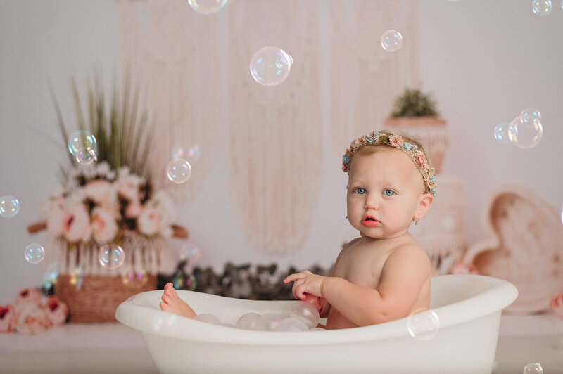 One year old baby girl milestone portrait in studiofloral tiara while sitting in a white bathtub surrounded by bubbles in front of a boho and floral backdrop