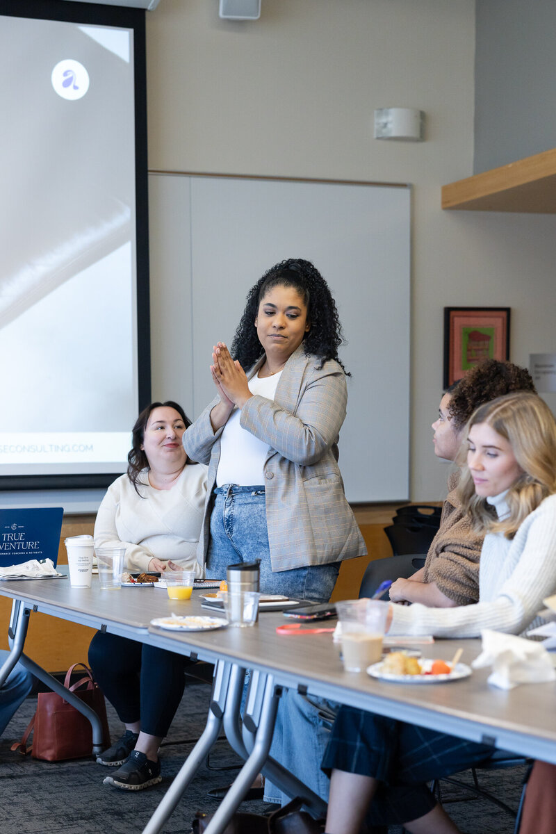 A group of people are seated closely together in a room at a networking event in Seattle, facing a speaker at the front. One woman in the foreground is holding open a notebook and writing with a purple pen. The chairs are quilted with a pinkish hue.