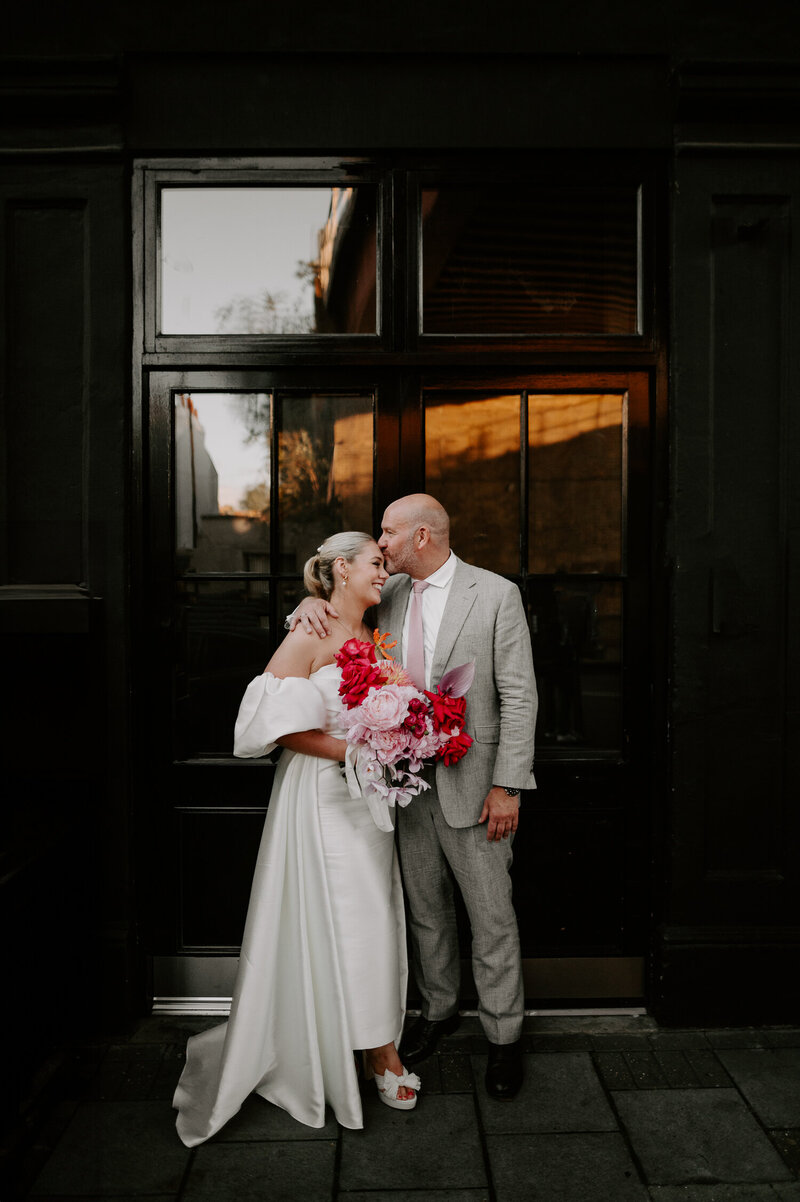 A groom kisses his bride on the forehead outside 100 Barrington in Brixton, London. The city wedding venue has a cool industrial black door.