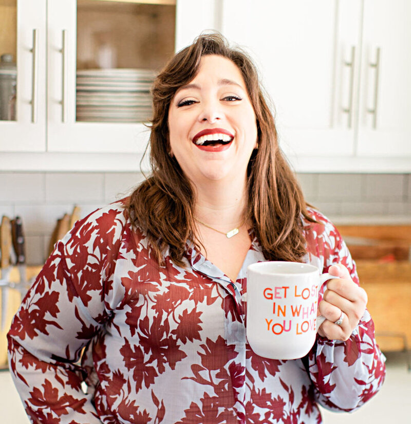 Renee Dalo laughs in floral blouse while holding coffee mug in kitchen