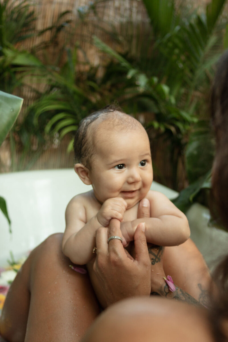 Baby smiling in a floral bath, held gently by a parent, captured during a creative San Diego newborn photography session