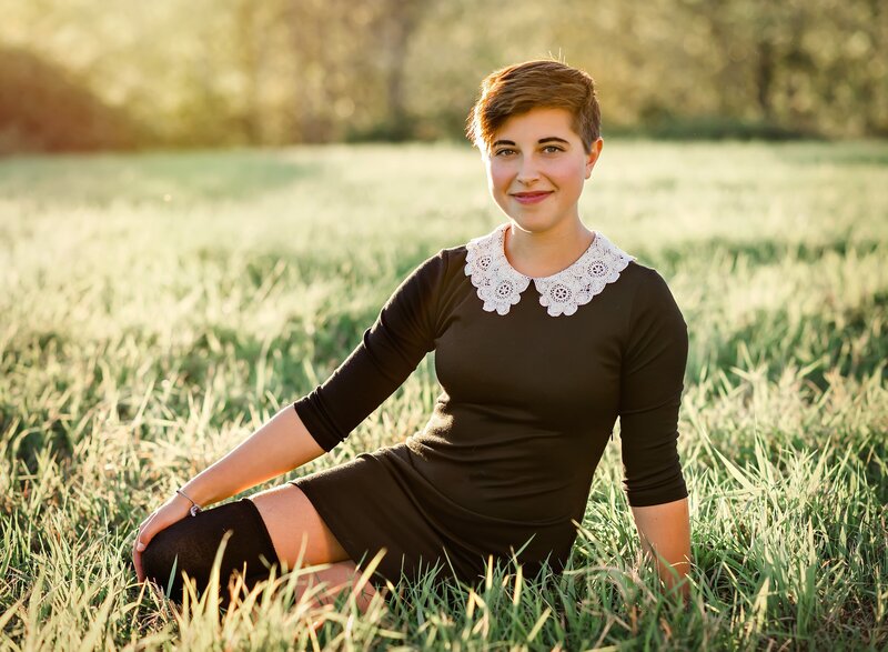 Teen girl in black dress and short hair smiles for senior portrait in a grassy field