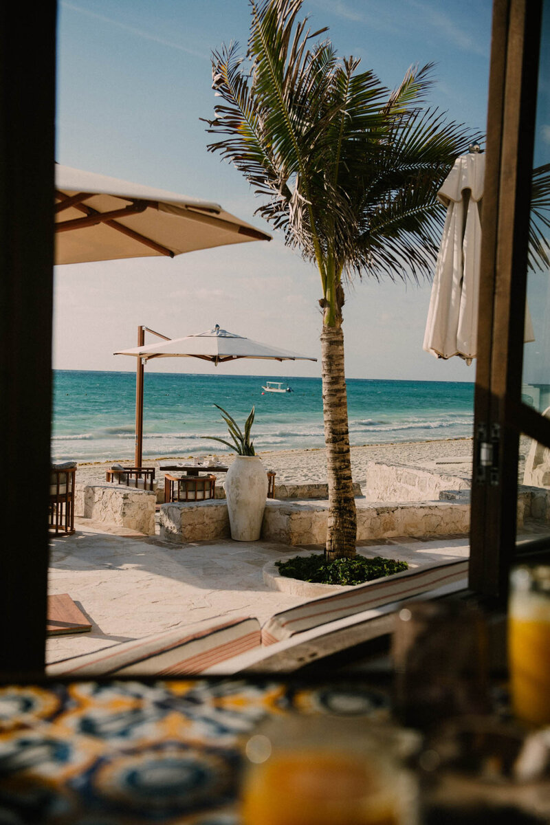 View from a window overlooking a beach with sandy shores. A palm tree and two outdoor tables with large umbrellas stand on a patio. The turquoise ocean and a distant boat are visible under a clear blue sky.