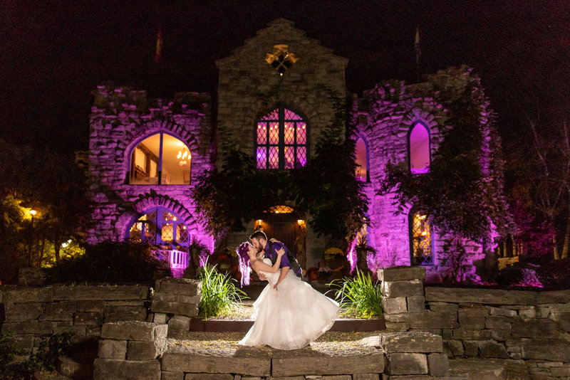 Groom dips bride in front of castle lit in purple