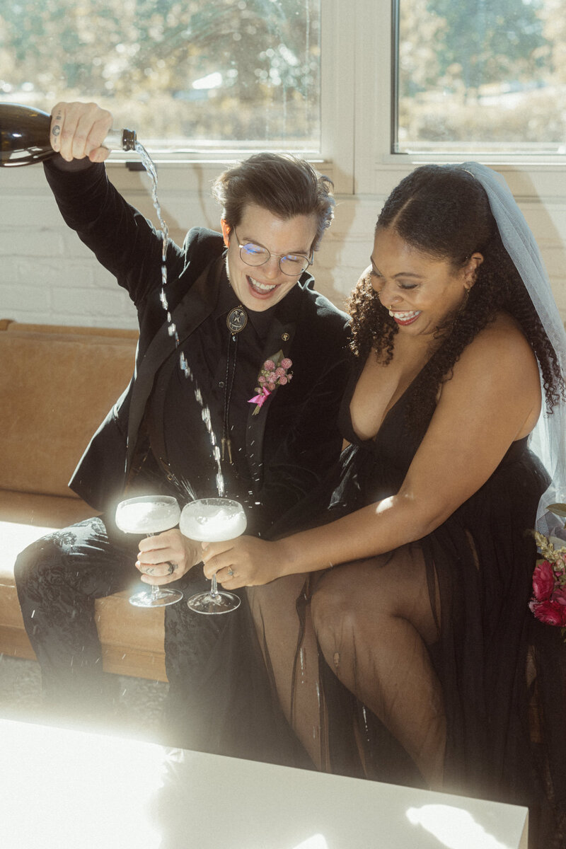 A queer couple sits next to each other on a sofa. One wears a black suit with a silver bolo tie, and the other wears a black dress with a grey veil. The partner in the suit pours champagne into their champagne coupe glasses.