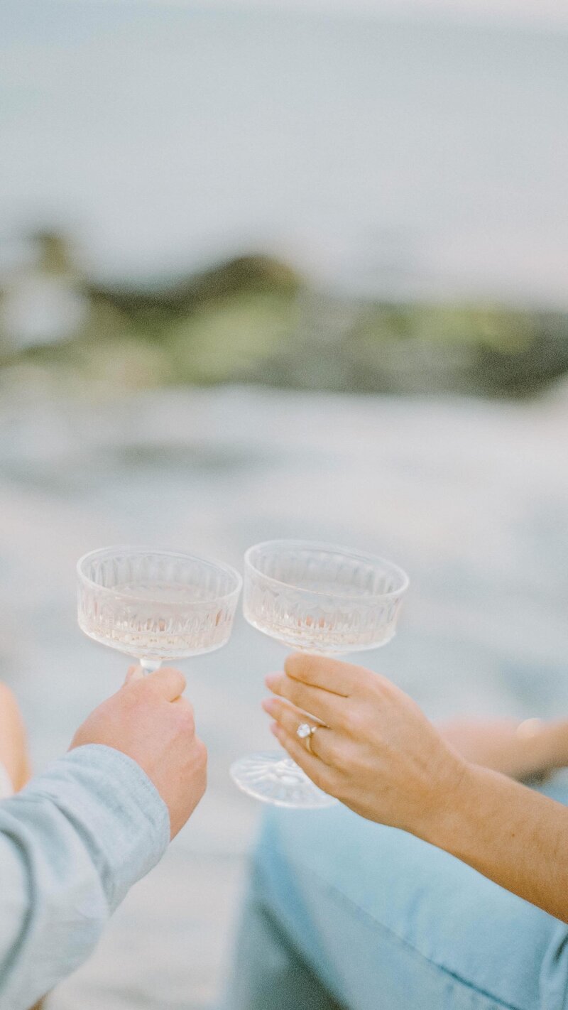 Wedding Photographer RI | Two people clink champagne glasses near a blurred beach scene. 