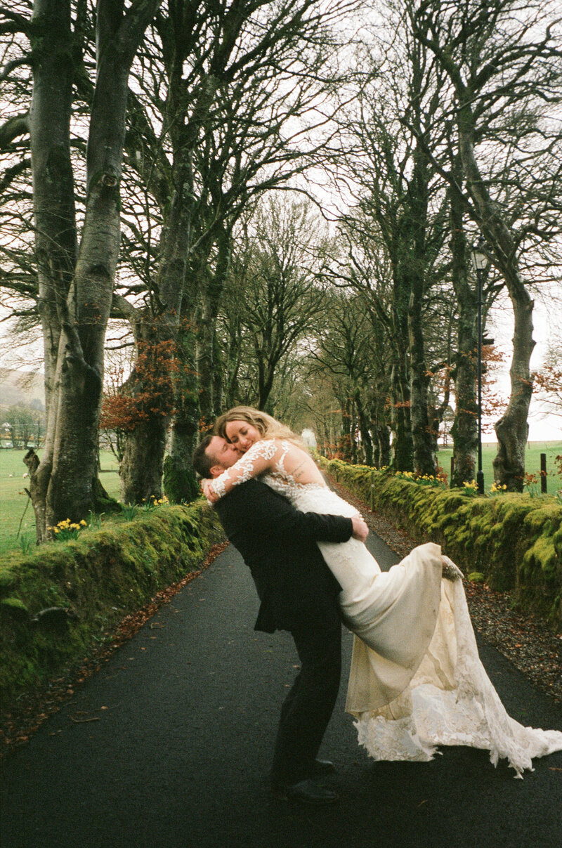 A groom playfully lifting his bride in a romantic embrace on a tree-lined avenue