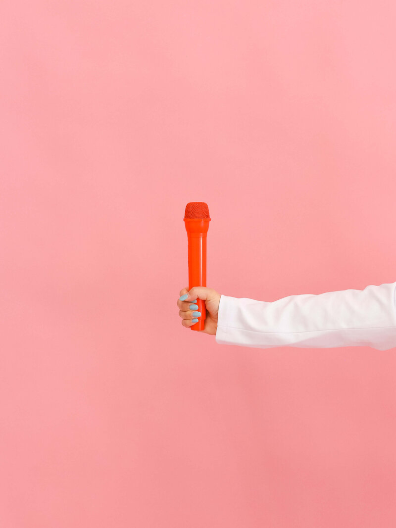 A person wearing a white sleeve holds an orange microphone against a plain pink background.