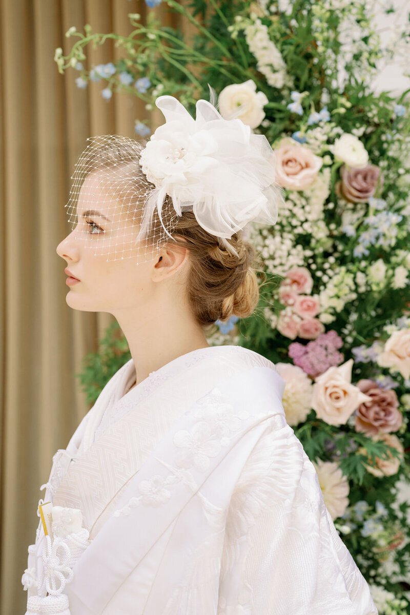 Bride wearing shiromuku (白無垢) wedding kimono in a traditional Japanese wedding ceremony with floral background.