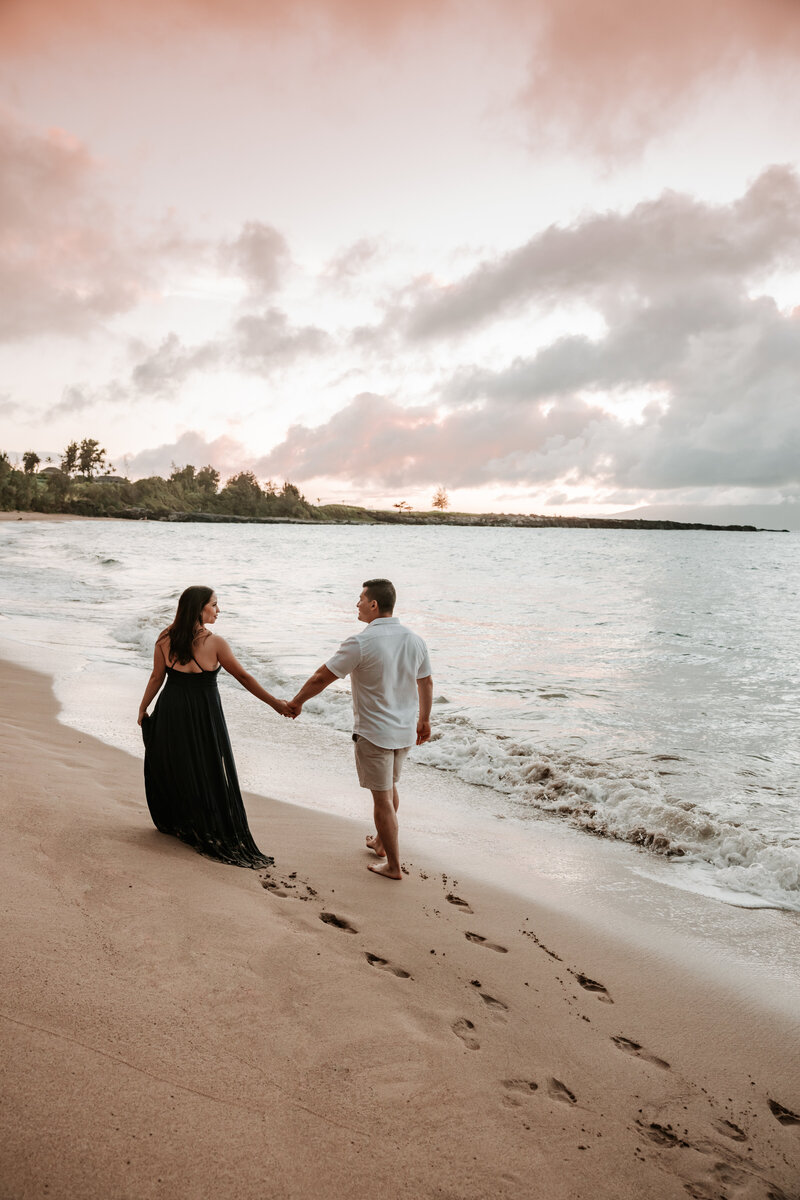 couple who is walking along the beach in Maui at sunset holding hands. she is in a beautiful long dark blue dress and he is in a neutral shirt and pants