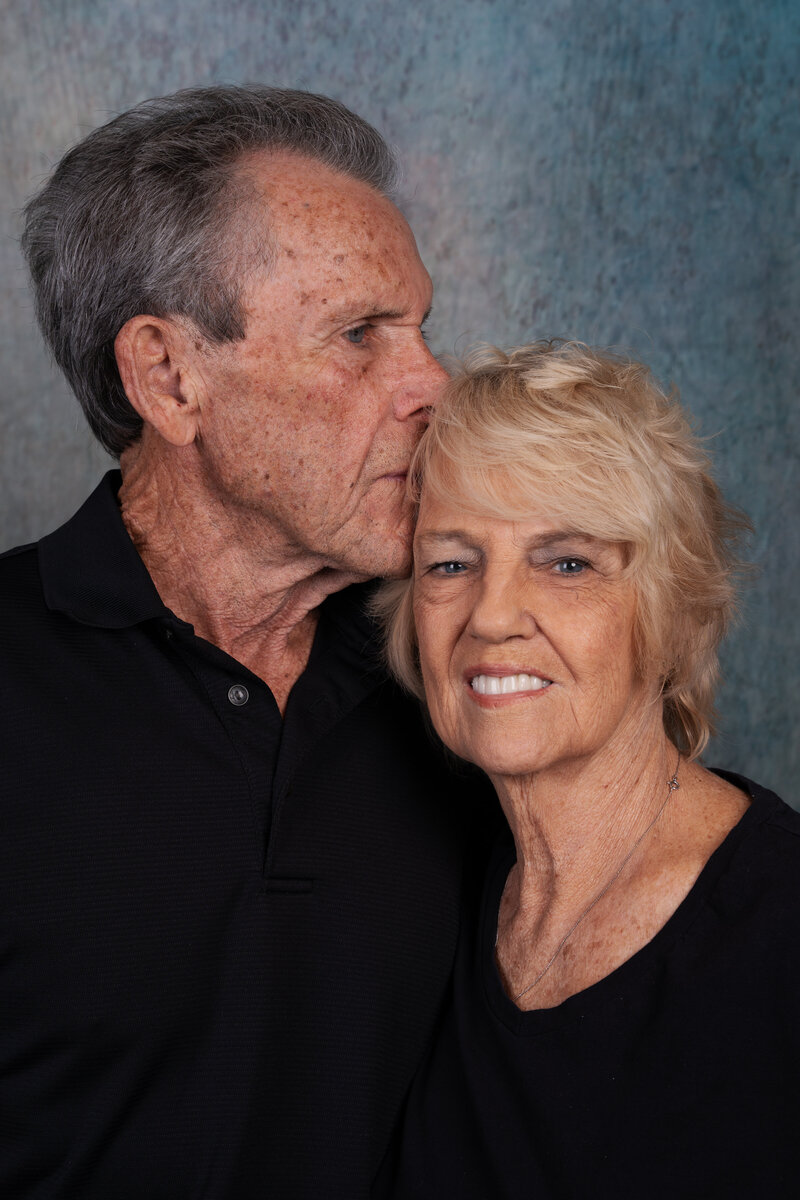a photo of an elder couple as he gives his wife a kiss on the head