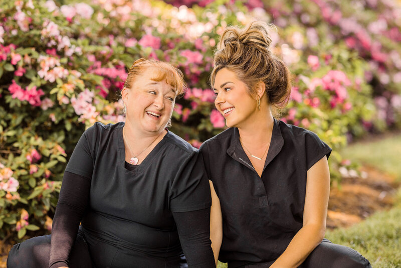two women wearing black scrubs sitting in front of azalea bushes smiling at each other