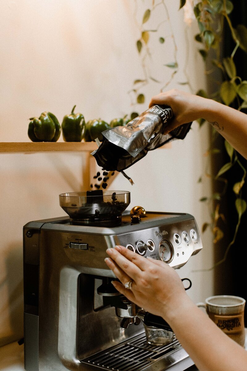 A person pours coffee beans into a silver espresso machine on a kitchen counter. Green bell peppers sit on a shelf above, while plants hang in the background—a scene perfect for anyone taking a white label branding course from home.