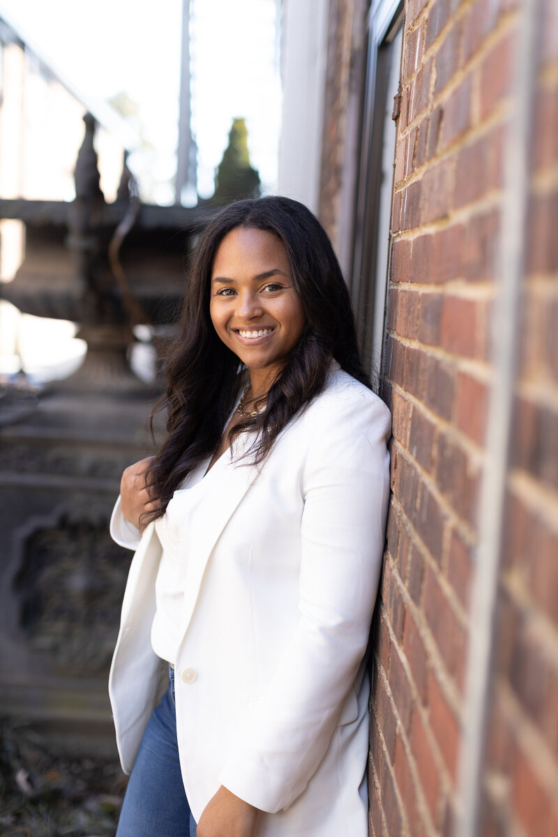 Niyani smiling, wearing a white blazer leaning against a brick wall.