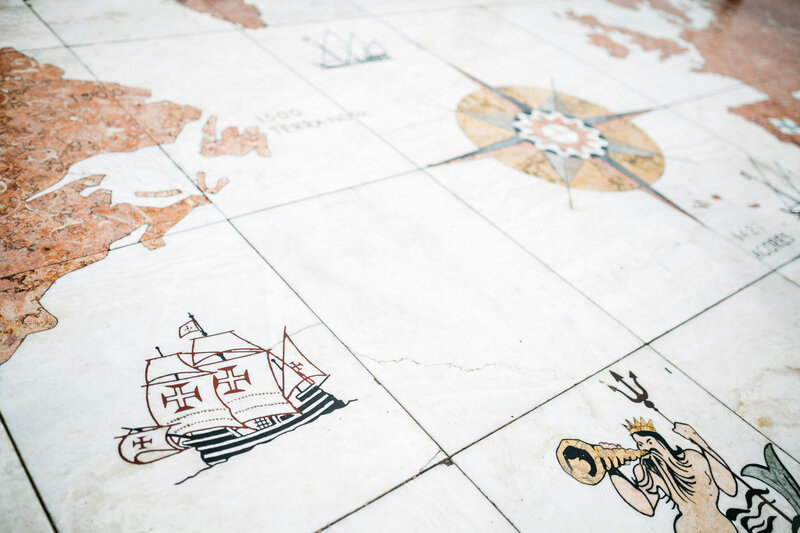 A close-up view of a large tiled map featureing historical ships with sails adorned with crosses, a compass rose, and a figure holding a trident and horn