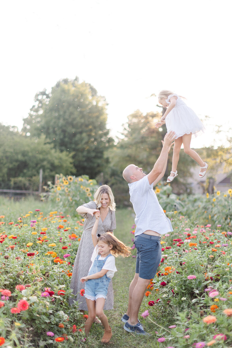 Dad throws his daughter in the air as mom twirls their other daughter behind them in a garden