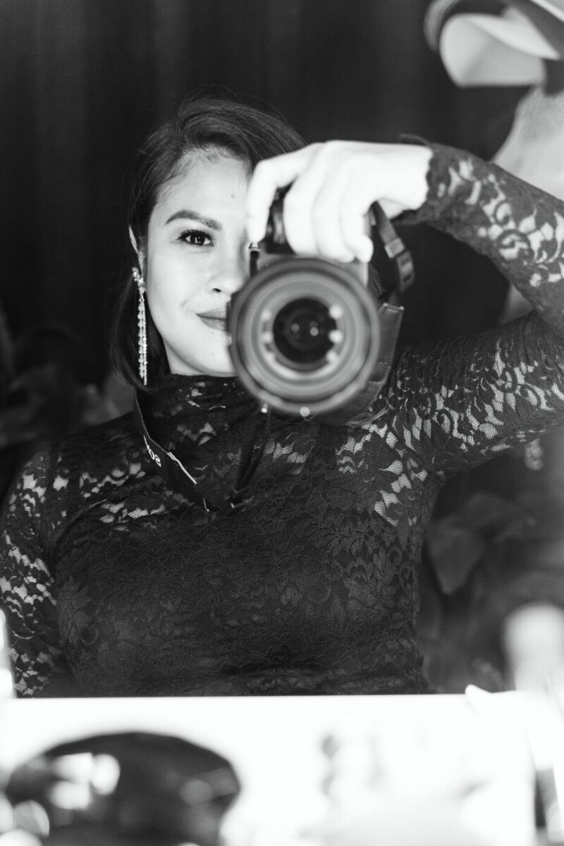 Black-and-white portrait of a smiling woman photographer wearing a lace dress and long dangling earrings, holding a professional camera to capture a shot.