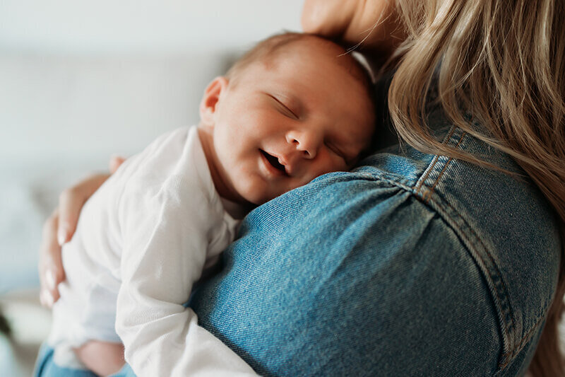 Newborn baby boy smiling while laying on mom's shoulder during an in home session