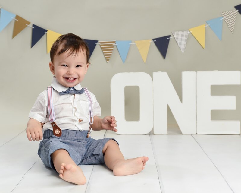 A baby boy in a white shirt and blue shorts smiling next to a "ONE" sign, in a pastel-themed cake smash setup with bunting in the background.