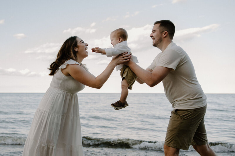 Family photography session at the beach at Lake Michigan, Milwaukee WI in front of the water, mom and dad are passing toddler son from dad to mom and they are all smiling