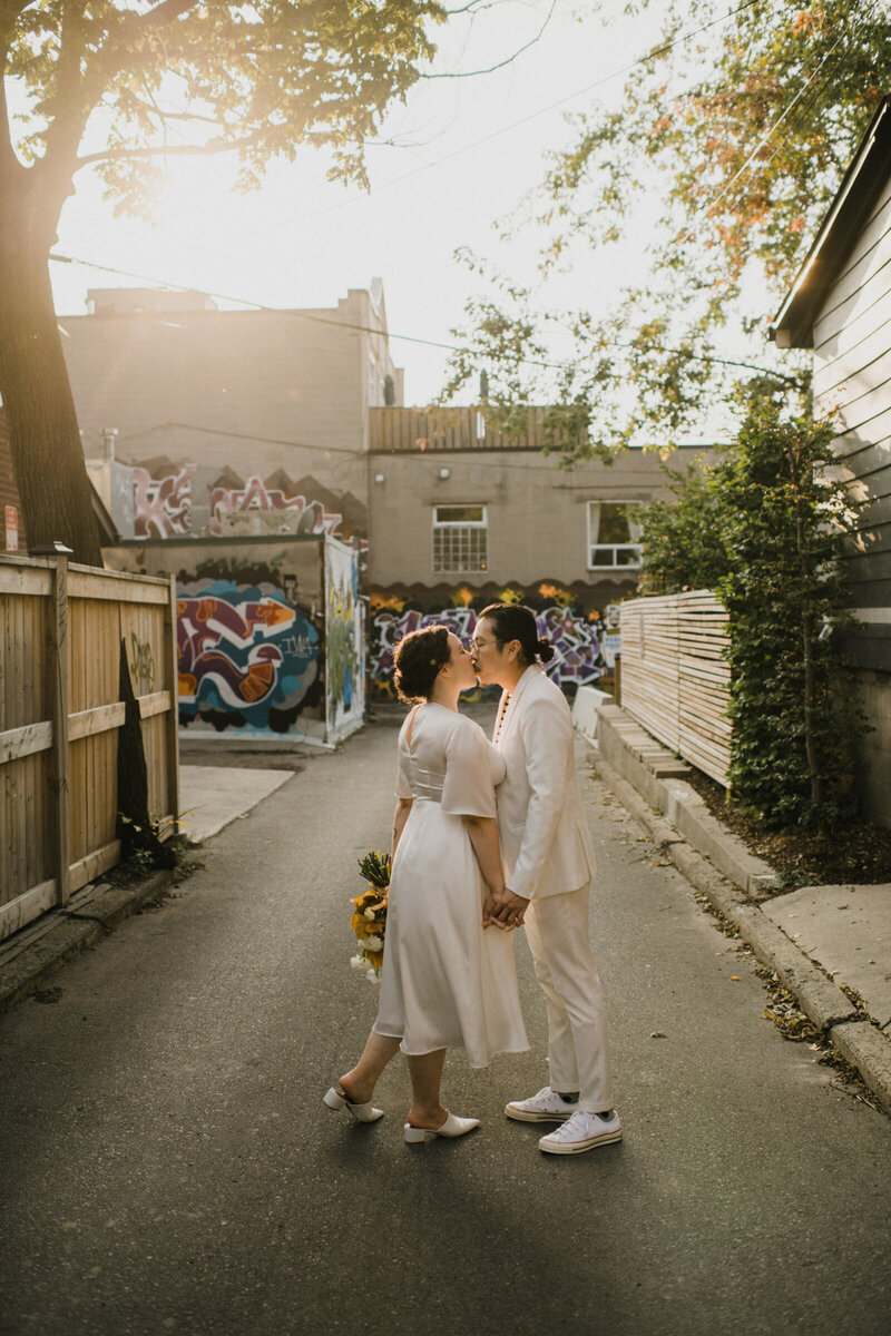 Queer wedding couple dressed in all white, kissing in alleyway in downtown Halifax.