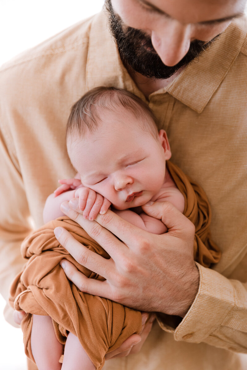 Close up of newborn baby in rust orange wrap against father's chest holding dad's hand with tiny fingers - professional newborn photography by Glean and Co Boise studio