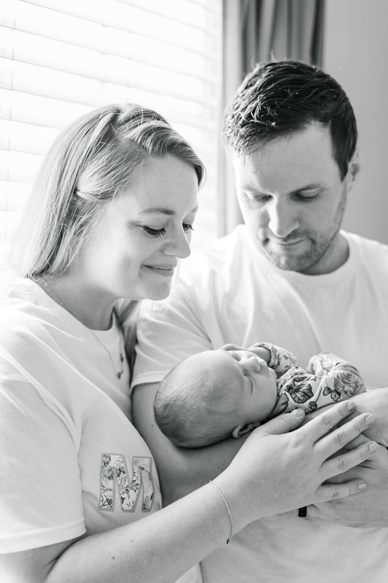 A couple stands in front of a window. The father holds their newborn baby while the mother wraps her arm around his arm. They both look at their baby with a soft smile while captured by a Round Rock newborn photographer.