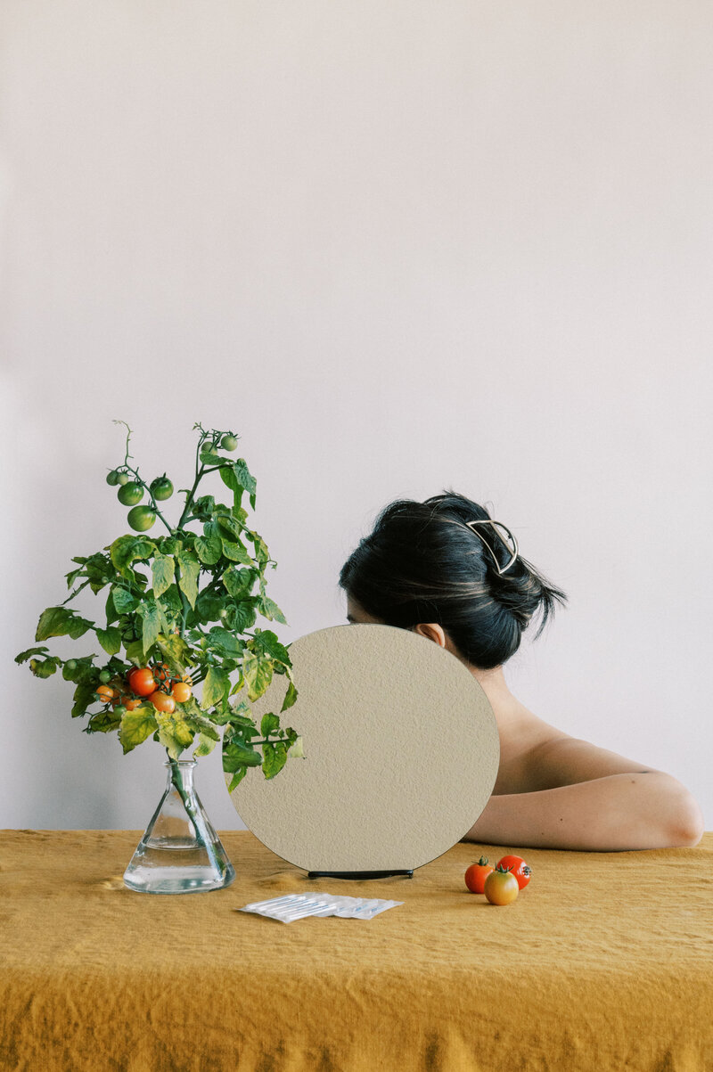 Woman with hair up sitting behind circular mirror beside vase with tomato plant