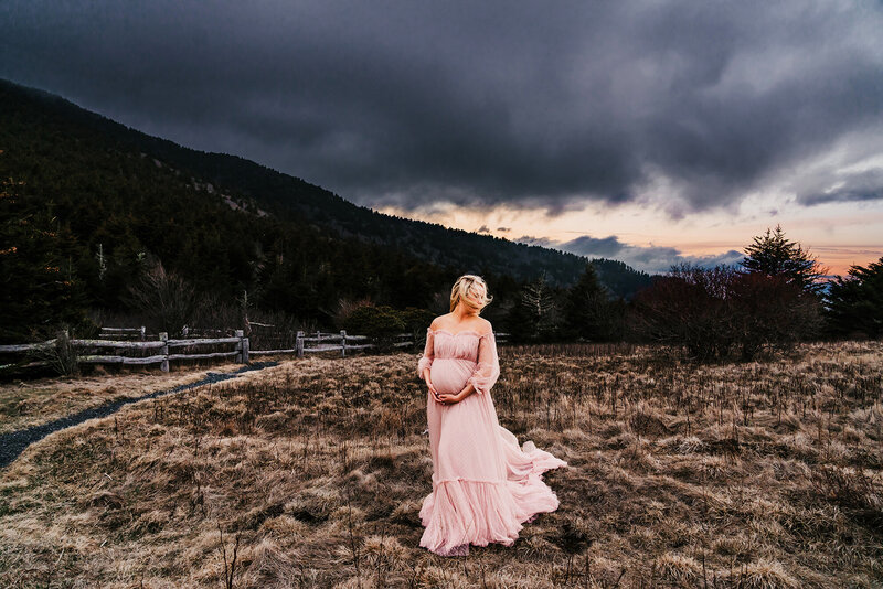 a pregnant mama in a pink gown holding her belly with the wind in her hair on a mountain