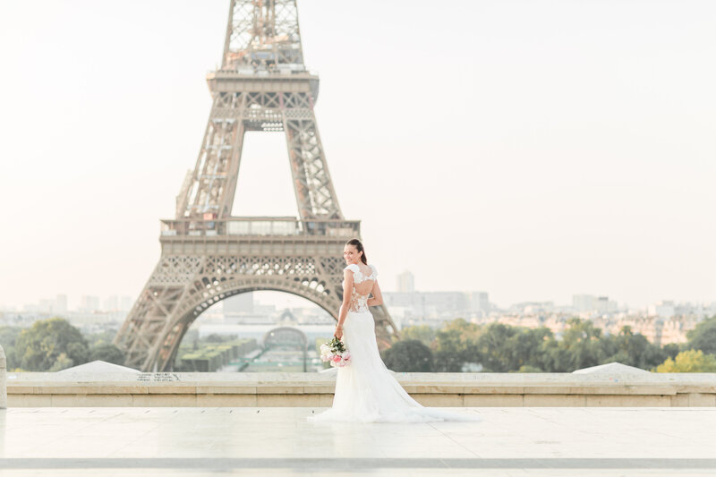 Bride poses at le Place du Trocadéro in Paris, France with the Eiffel Tower clearly in behind her. She is facing the Eiffel tower and is looking over her shoulder and smiling at the camera. Captured by best Paris destination wedding photographer Lia Rose Weddings