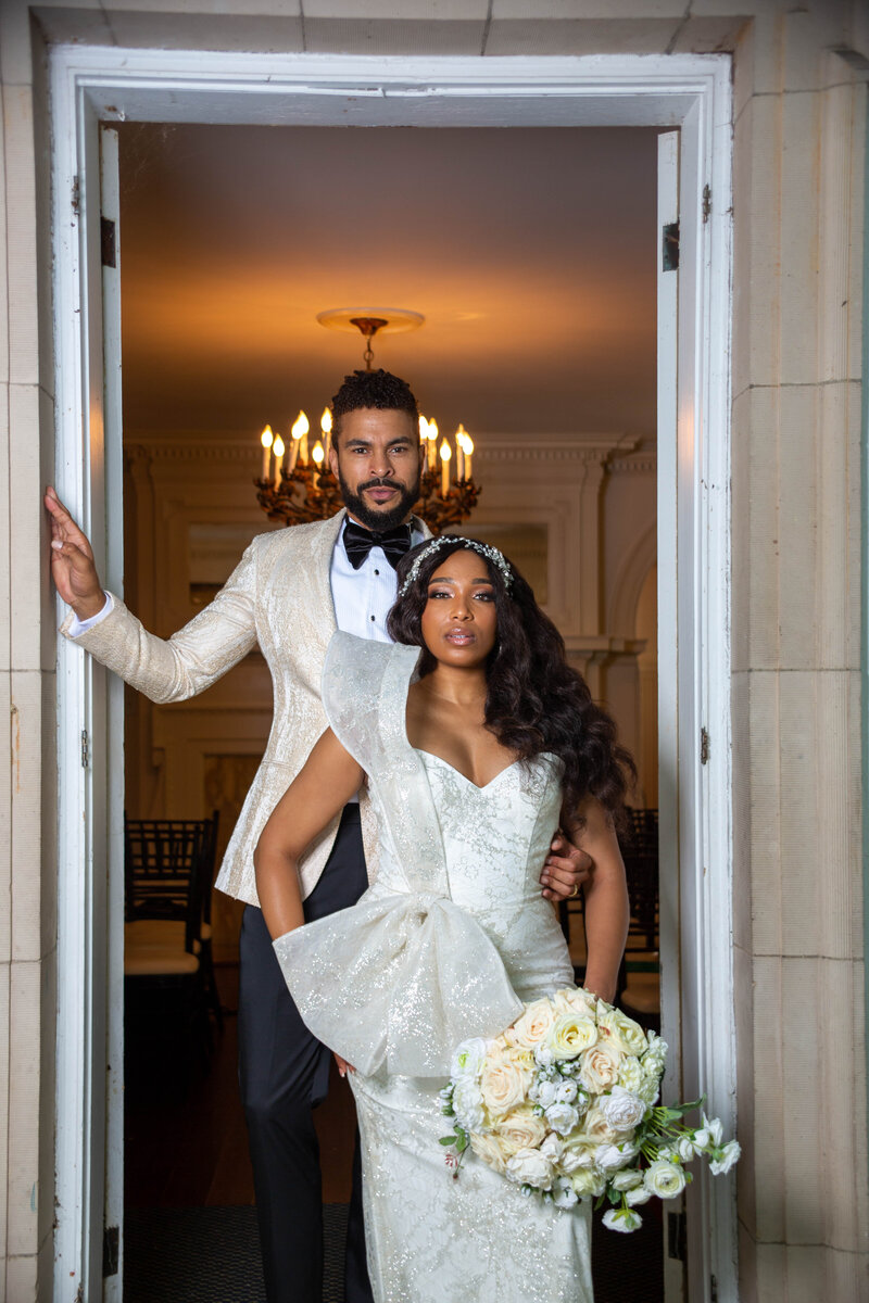 Couple stand in doorway dressed in white attire.