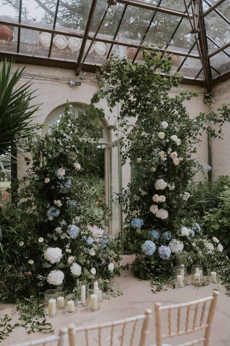 A bride in a white wedding dress with puff sleeves stands in front of a large, luxurious and statement wedding ceremony backdrop made up of colourful flowers and wild foliage.
