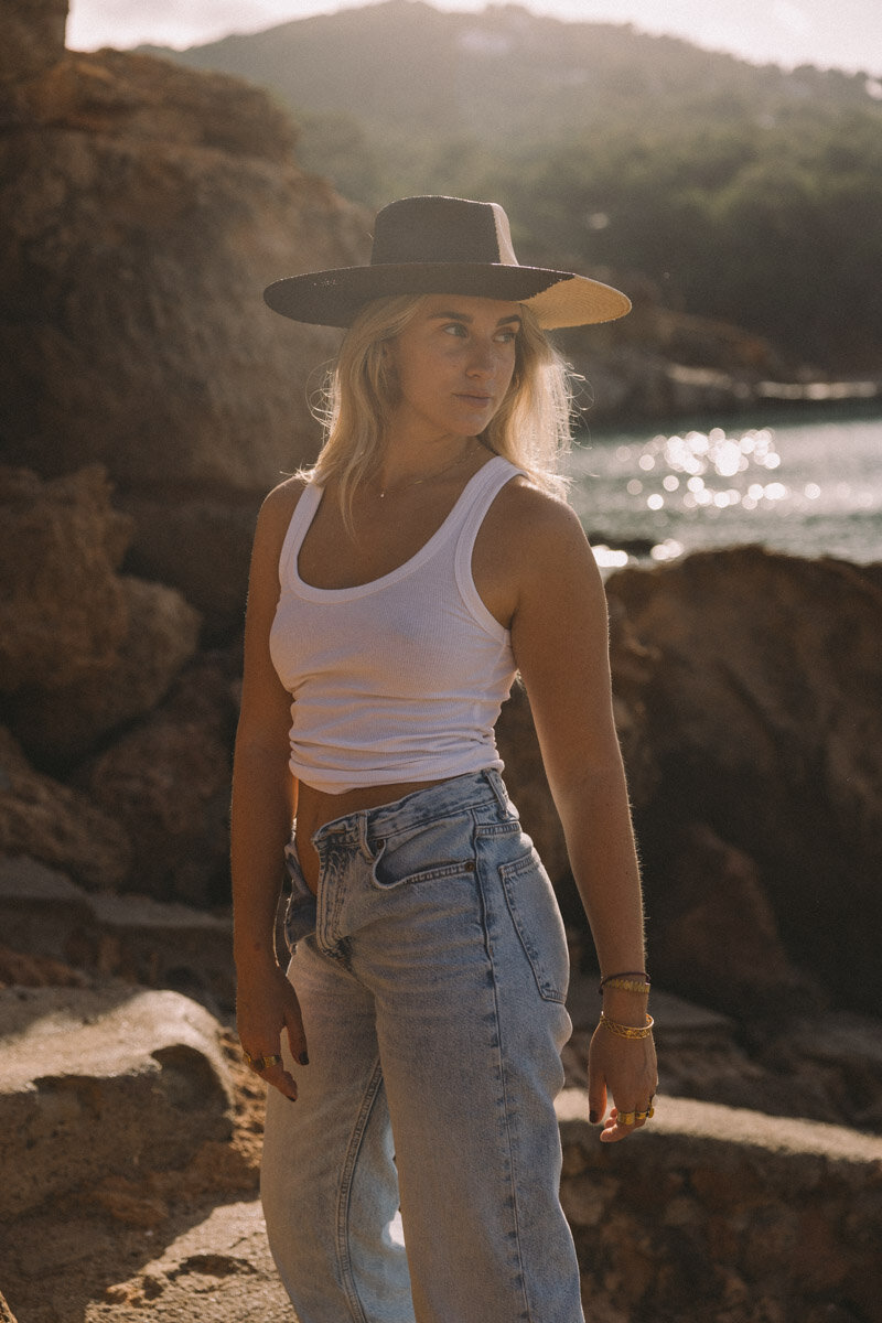A woman in cowgirl hat in white top and jeans is posing on the beach.