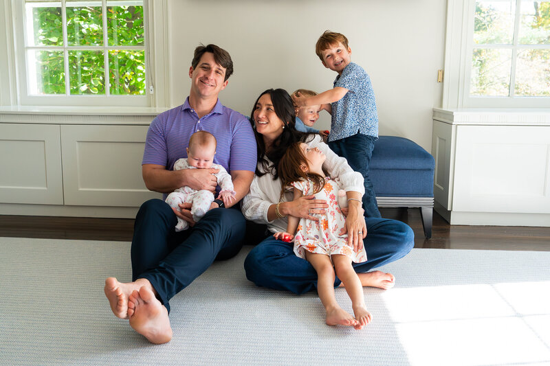 Family with three small kids and a baby sitting together on bedroom floor.