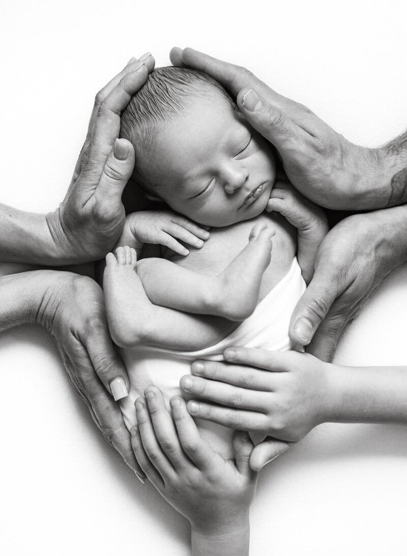 Black and white photo of newborn boy sleeping with mom, dad, and sibling hands wrapped around curled baby.