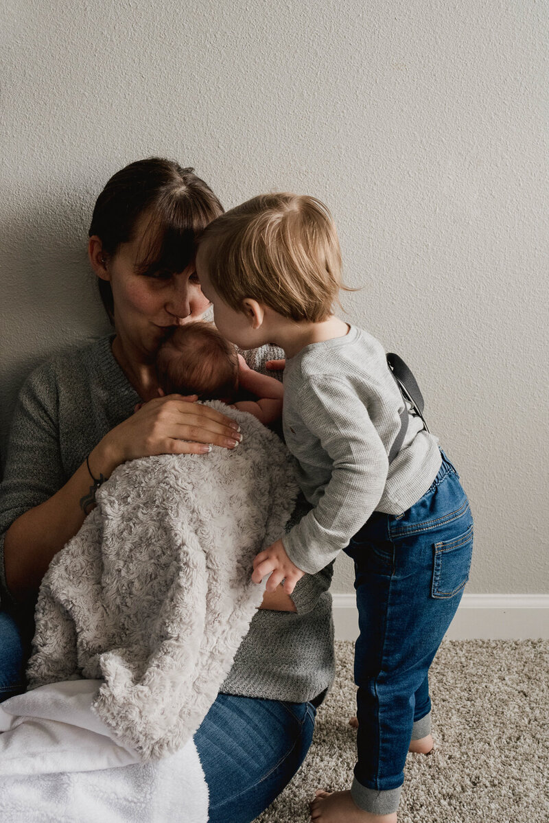 Mom resting on the floor, sitting against the wall holding newborn son under a gray blanket. The sibling is leaned over giving his baby brother a kiss on the back of his head.