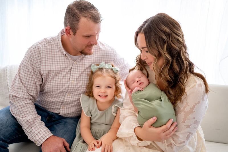 Bright and joyful newborn family portrait featuring mom, dad, big sister, and baby wrapped in sage green, captured in a light-filled Waukesha photo studio near Brookfield and Lake Country, WI.