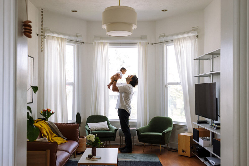 dad standing in living room in front of windows and holding baby up to play airplane
