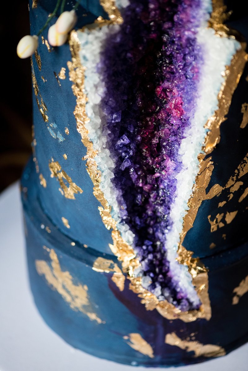 3 tier navy cake with gold foil detailing and purple agate design cut into the side, toped with pink and white orchids