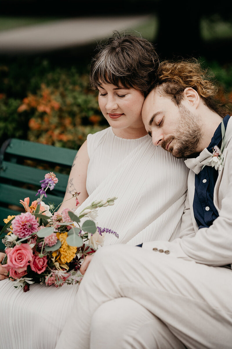 Newlyweds share a quiet moment on a park bench with a wildflower bouquet.
