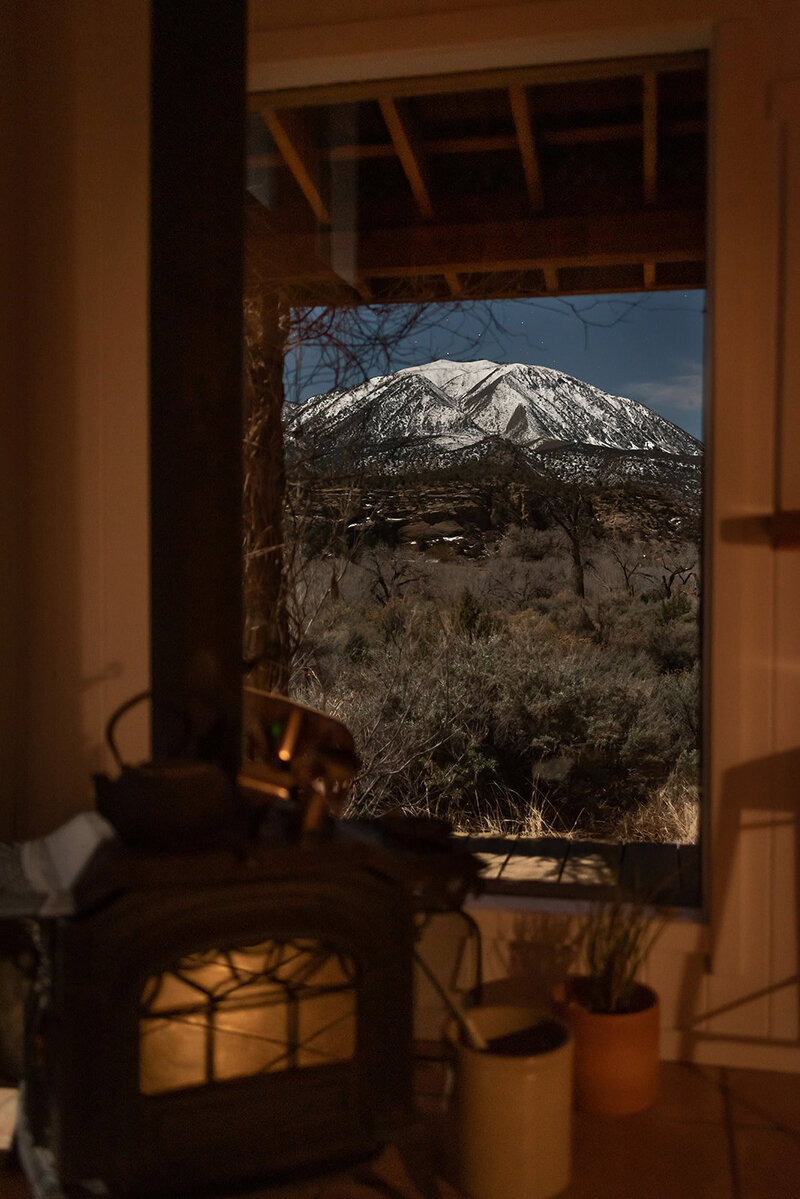 a luxury elopement photographer during a remote Colorado elopement wedding captures a night scene of a mountain capped in snow glowing in the moonlight  out the window of a cabin where a cast iron wood stove has a gentle warm glow in the glass door of it's belly.  